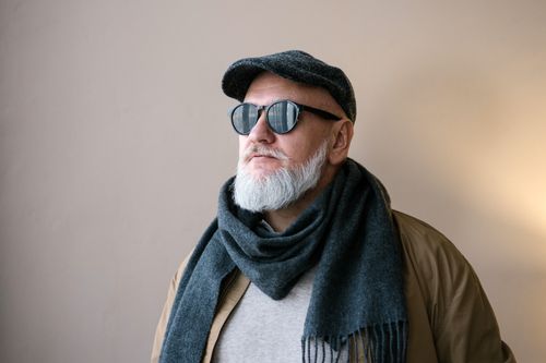 Free Portrait of a stylish elderly man wearing sunglasses, hat, and scarf in an indoor setting. Stock Photo