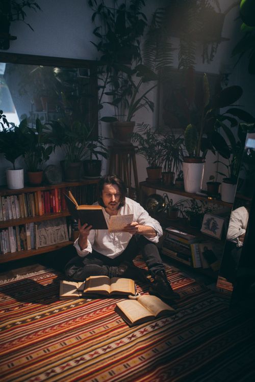 Free A man sits reading books surrounded by decorative plants in a cozy room, creating a scholarly atmosphere. Stock Photo
