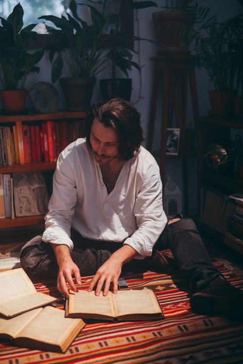 Free A man in a white shirt sits on a rug, reading vintage books in a cozy home library setting. Stock Photo
