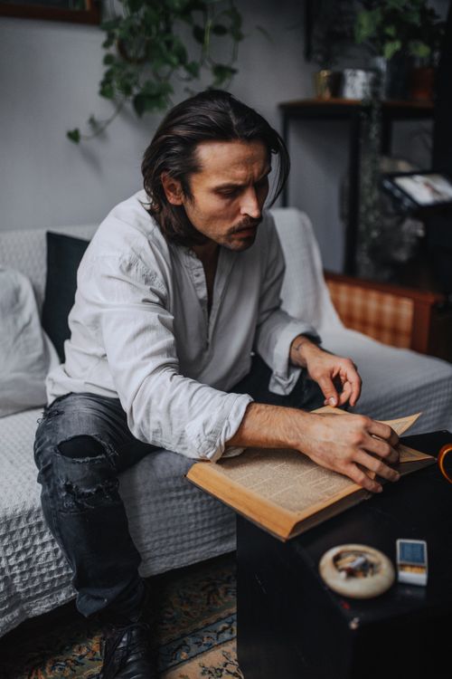 Free A man with long hair sitting on a couch, focused on reading a book in a cozy home setting. Stock Photo