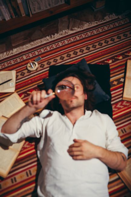 A man lies on a striped rug, holding a magnifying glass, surrounded by books. A man lies on a striped rug, holding a magnifying glass, surrounded by books.