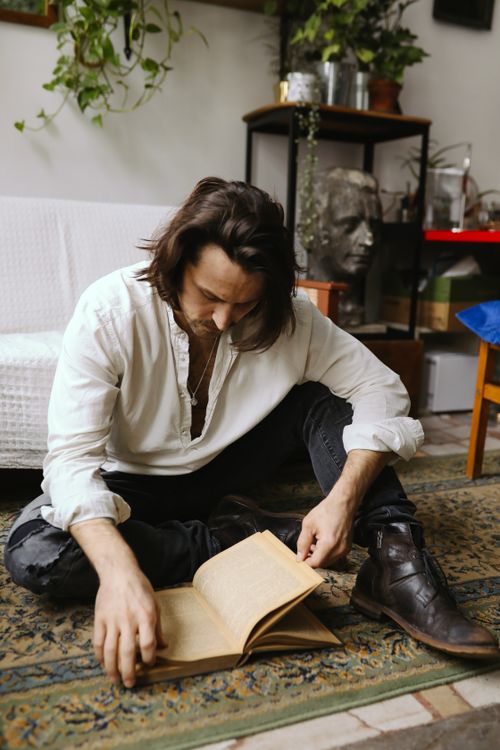 Free Thoughtful man reading a book while sitting on a patterned rug in a cozy indoor setting. Stock Photo