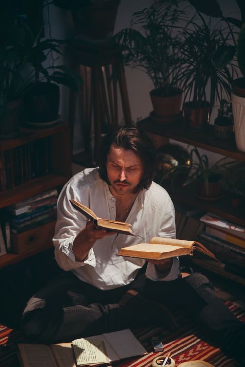 Free A man sits in a cozy room surrounded by books and plants, deeply engaged in reading. Stock Photo