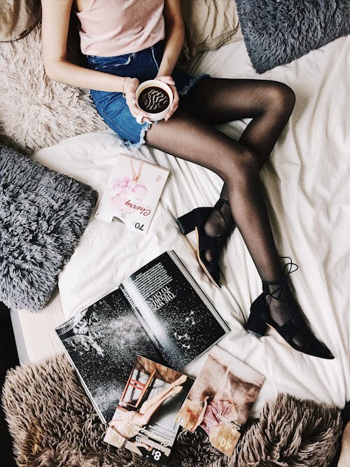 Flatlay фотография женщины, держащей белую кружку с черной жидкостью, лежащей на кровати в окружении меховых подушек и журналов