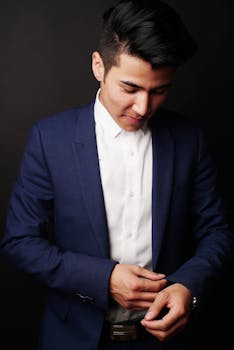 Confident young man in a tailored blue suit buttoning his jacket against a black studio background.