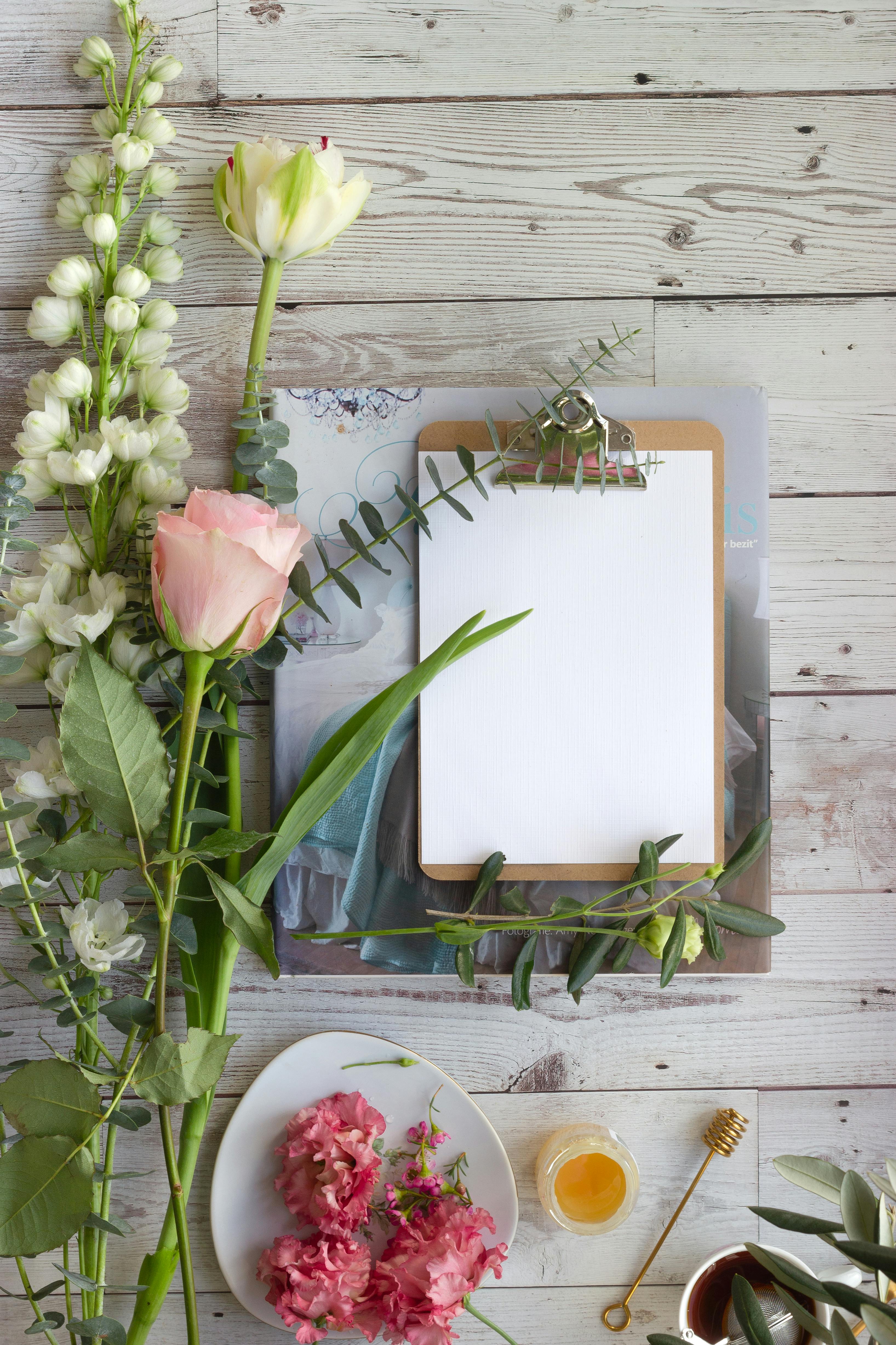 Flat lay of flowers, clipboard, and honey dipper on rustic wood background, ideal for creative designs.