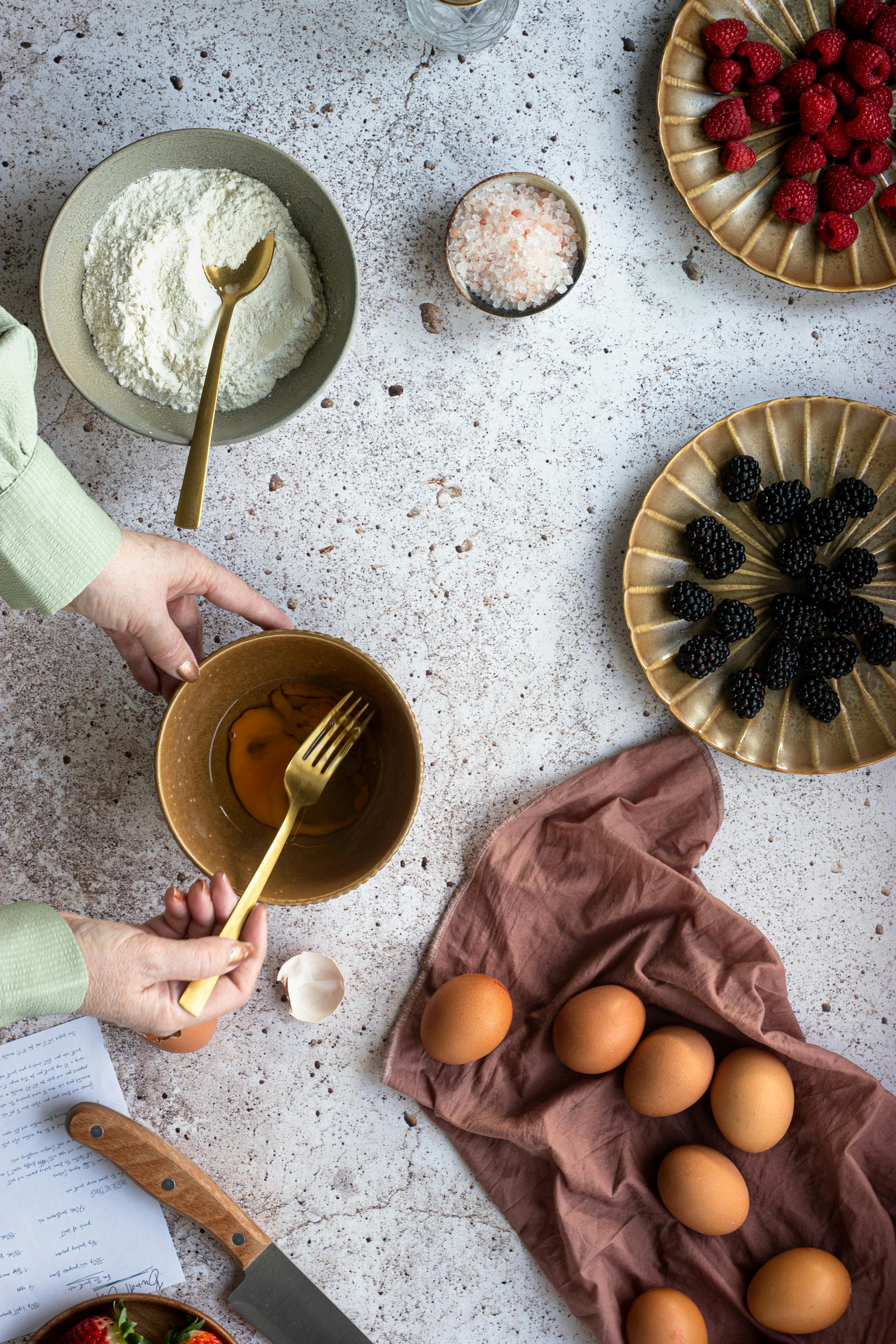 Overhead shot of food preparation with eggs, flour, berries, and mixing ingredients on a kitchen counter.