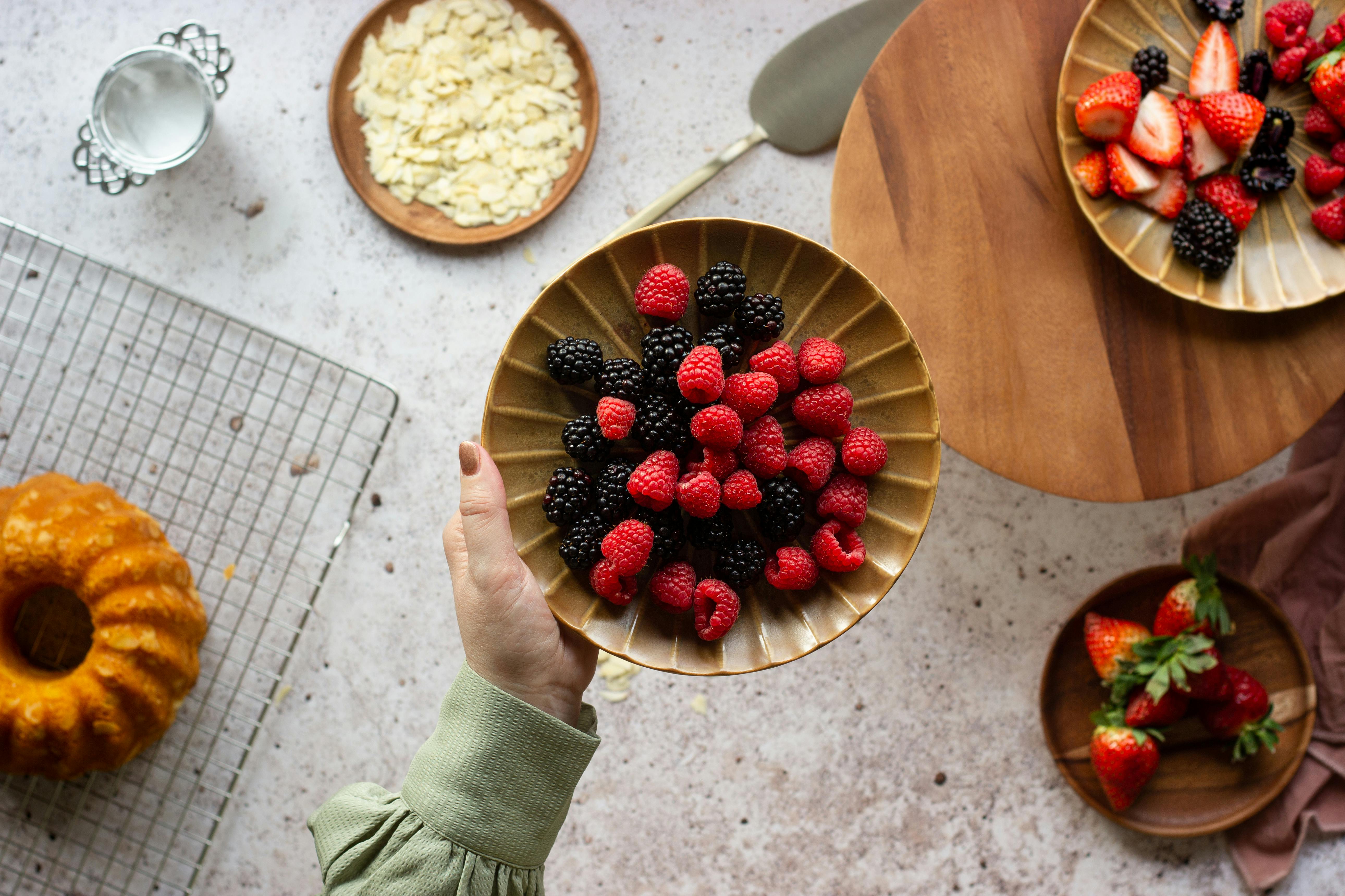 Overhead view of hand holding a plate of fresh blackberries and raspberries alongside dessert and sliced almonds.