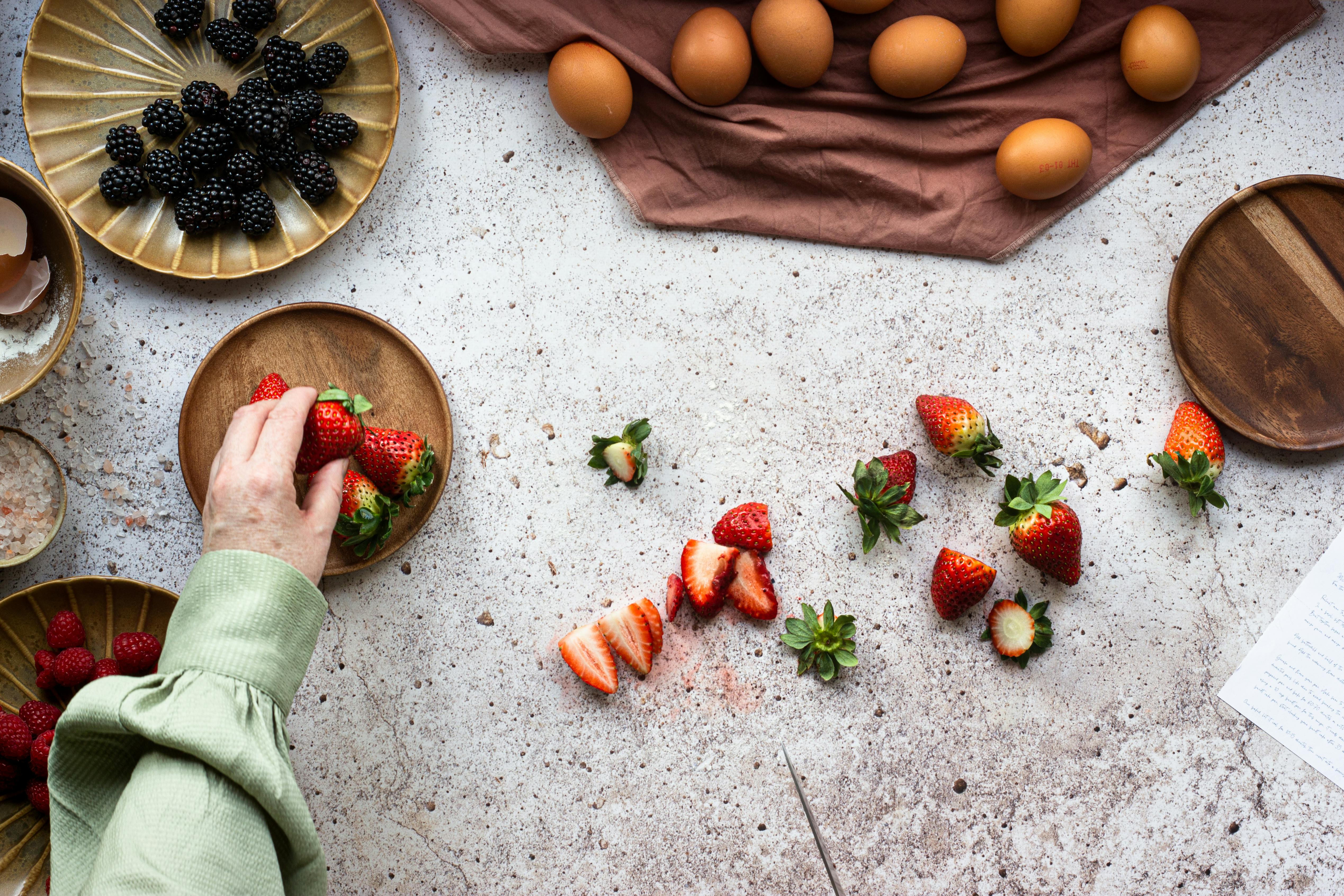 Overhead view of strawberries, blackberries, and eggs on a kitchen counter, highlighting food preparation.