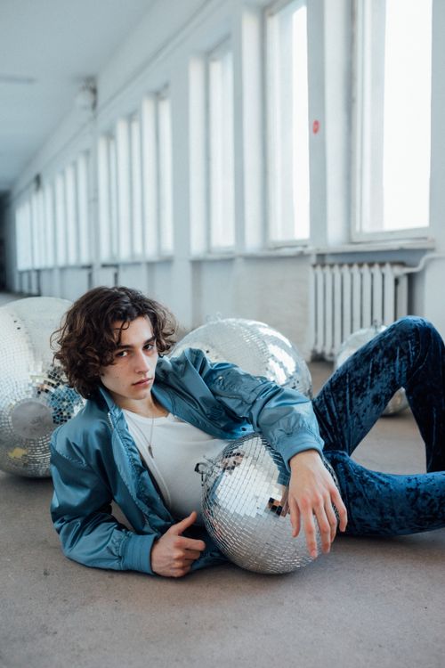 Free A fashionable adult male in a blue jacket poses with disco balls in a brightly lit hallway. Stock Photo