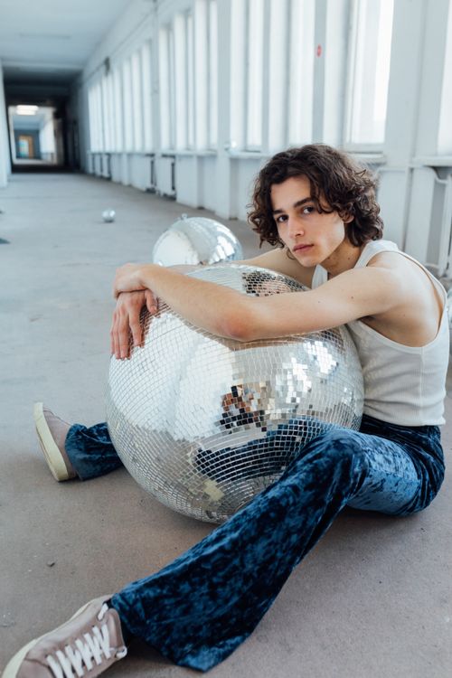 Free A young adult in a white tank top and blue jeans sits with a disco ball in a bright hallway. Stock Photo