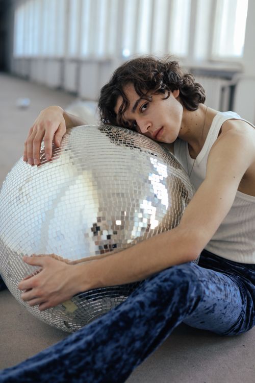 Free A young adult with curly hair embraces a disco ball in a bright hallway, wearing a white tank top and blue velvet pants. Stock Photo