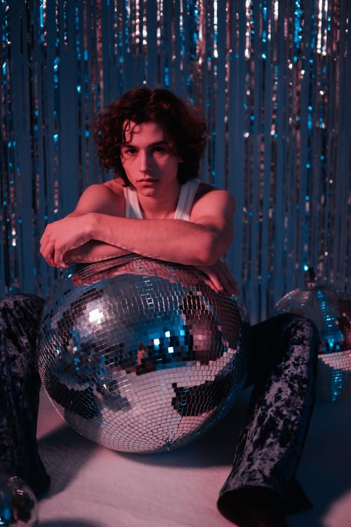 Free A young man sitting behind a disco ball with a retro foil curtain backdrop. Stock Photo