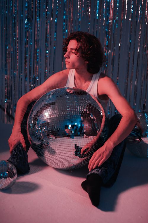 Free A young man sitting with a large disco ball in a vibrant retro-themed setting. Stock Photo