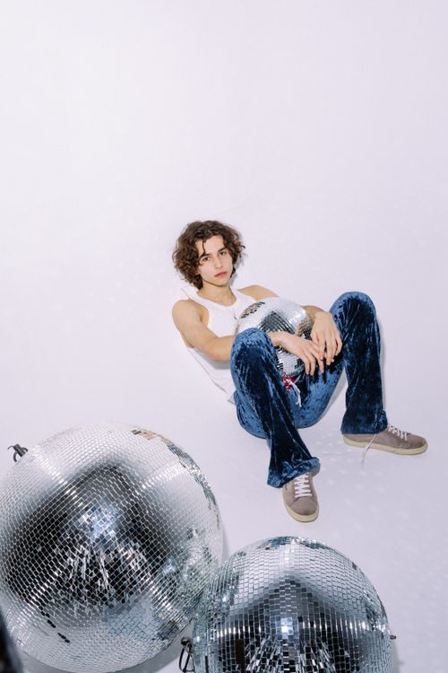 Free Portrait of a young man sitting with disco balls, wearing blue pants and white tank top. Stock Photo