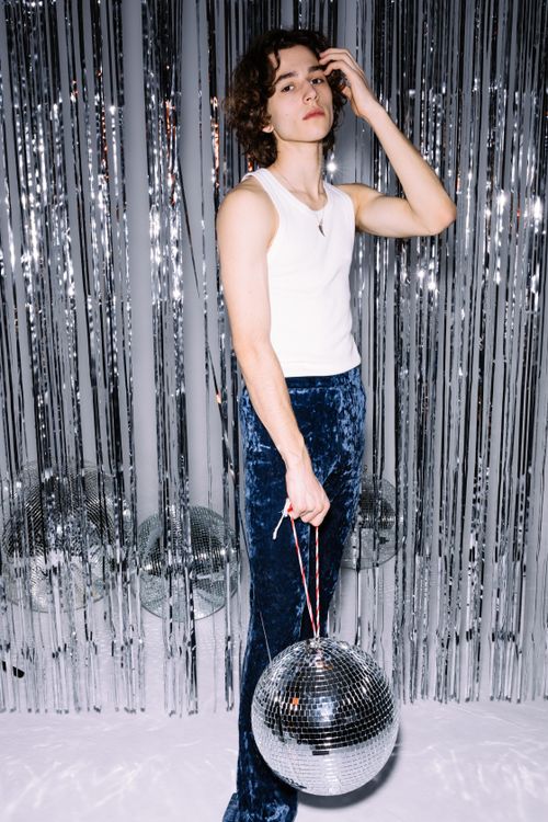 Free A young adult in a white tank top holds a disco ball amid metallic fringe. Retro theme. Stock Photo