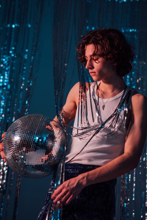 Free A stylish young man with a disco ball against a foil curtain backdrop, evoking retro vibes. Stock Photo
