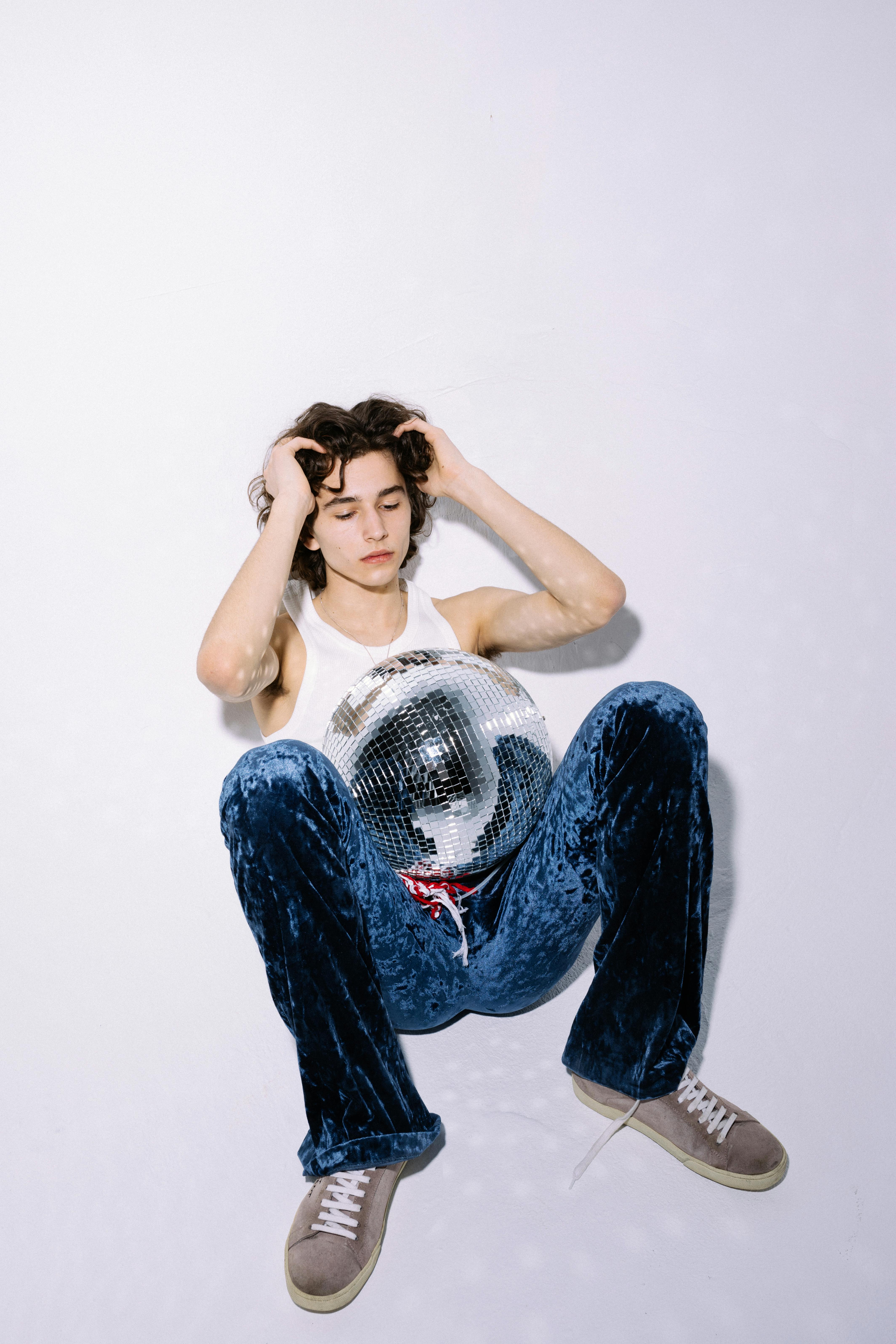 a man in white tank top and blue denim jeans sitting on floor with disco ball
