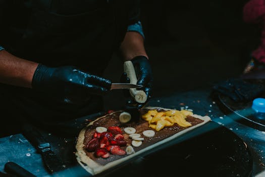 Chef slicing fruits for a delicious crepe with strawberries, bananas, and mango. Perfect street food delight.