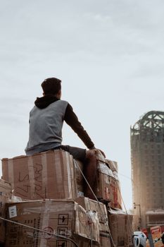 A man sits atop a stack of cardboard boxes, overlooking a cityscape against the sky.