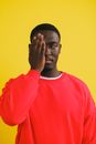 Calm African American male in red sweatshirt covering eye with hand and looking away against yellow background