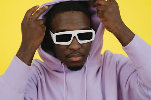 Trendy African American male in stylish white sunglasses putting on hood and looking at camera against yellow background
