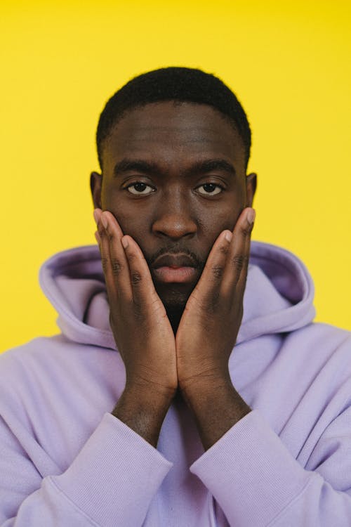 Free Calm African American male in purple hoodie touching cheeks with hands and looking at camera against yellow background Stock Photo