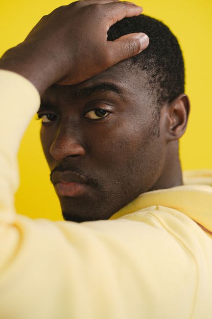 Serious African American male in hoodie touching head and looking at camera against yellow background