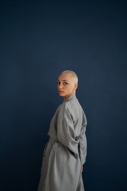 Back view unemotional bald female wearing gray robe standing with hands behind back and looking at camera against dark wall