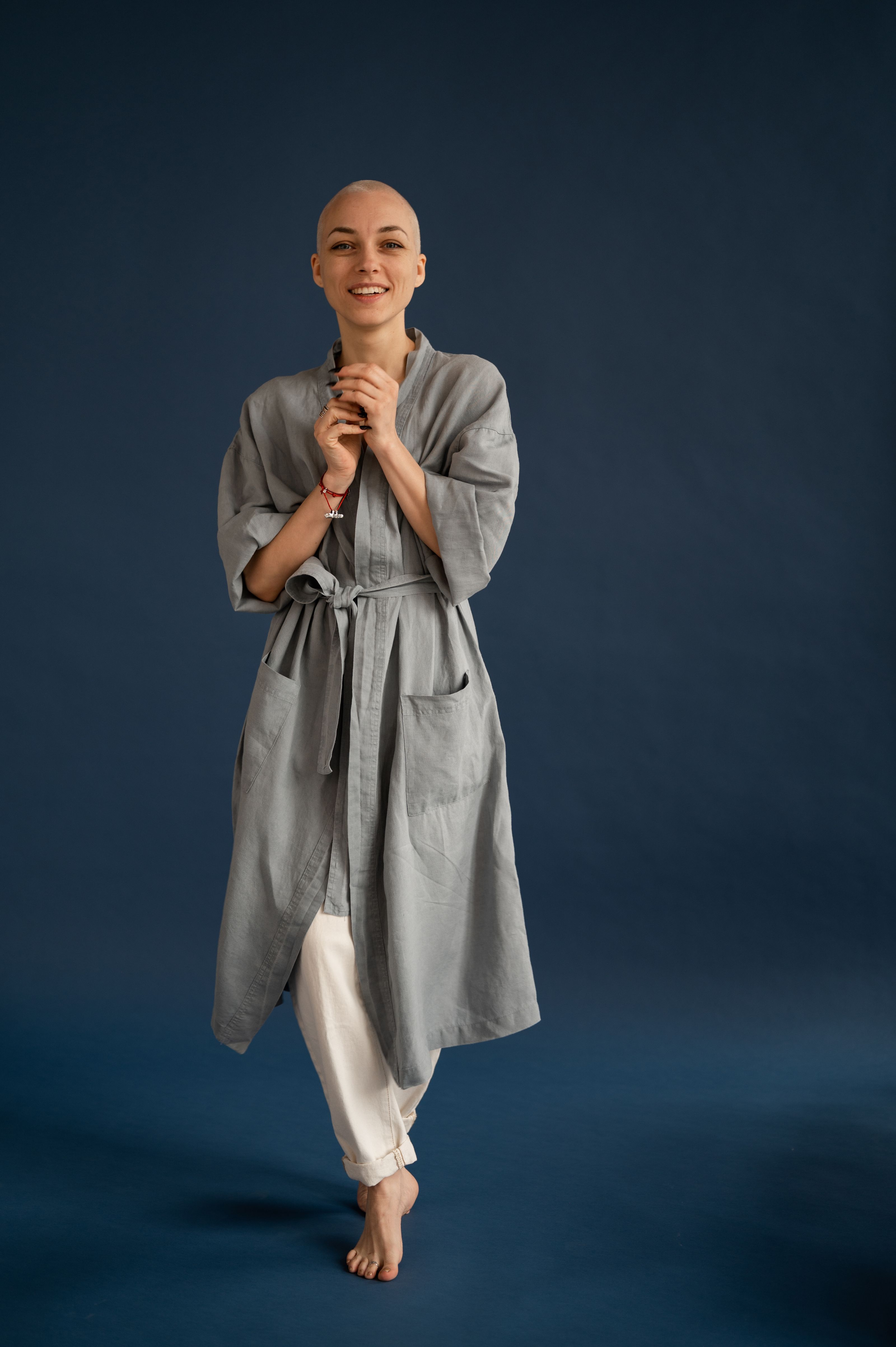 Mulher jovem e careca sorrindo com confiança em um estúdio, usando um roupão cinza elegante contra um fundo escuro.