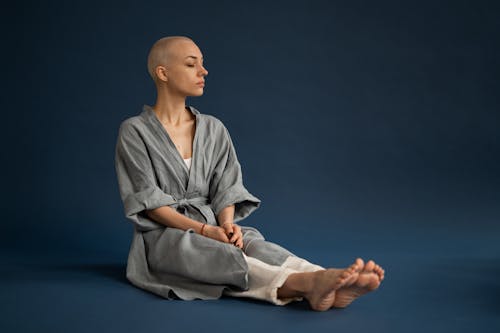 Full body thoughtful bald female wearing gray robe sitting with eyes closed on floor in dark photo studio