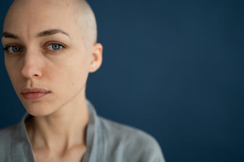 Crop wistful bald female in gray wear looking at camera thoughtfully against dark blue wall