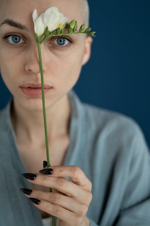 Free Crop unemotional bald female in gray robe touching face with white delicate flower and looking at camera against dark background Stock Photo