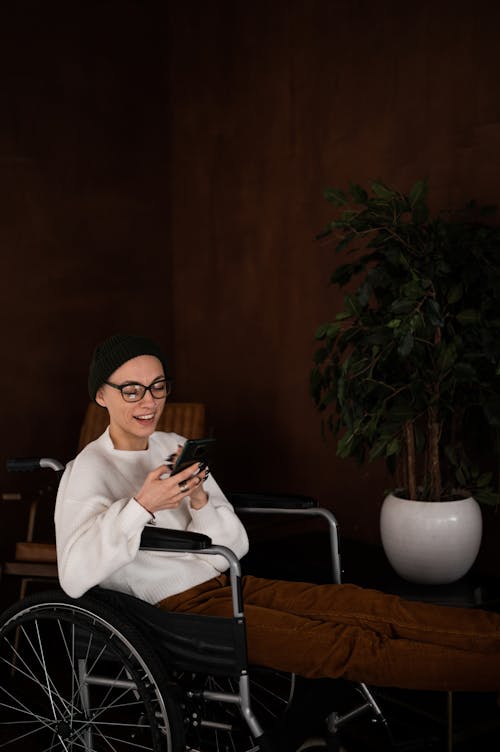 Free Side view of positive young female with disability in casual clothes and hat smiling and messaging on mobile phone while sitting in wheelchair Stock Photo