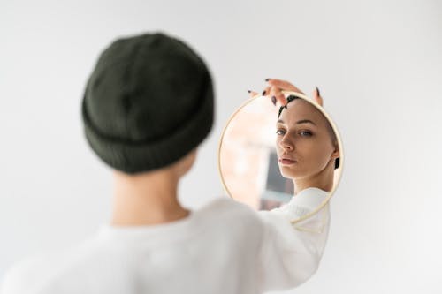 Free Back view of self assured young female in casual clothes and hat looking in round cosmetic mirror against white background Stock Photo