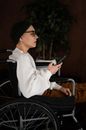 Side view of young concentrated female millennial in casual clothes and hat on bald head sitting in wheelchair with smartphone in hand and looking away thoughtfully