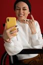 Crop optimistic female patient in casual clothes and hat smiling and showing sign of horns while having video call via smartphone sitting in wheelchair against red background