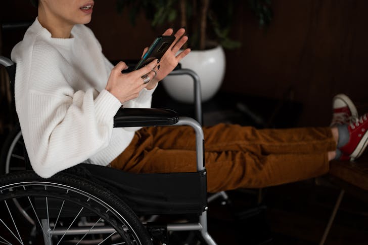 Side view of crop unrecognizable female millennial with disability sitting in wheelchair and browsing mobile phone