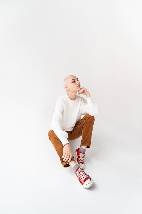 Full body of young thoughtful female in casual clothes with shaved head sitting on white floor in light studio with hand at chin