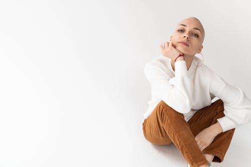 Thoughtful female with short blond hair in stylish clothes leaning on hand and sitting on floor in white studio