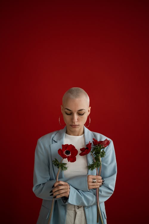 Pensive woman with short hair in stylish clothes standing with bright red poppies in studio against red background