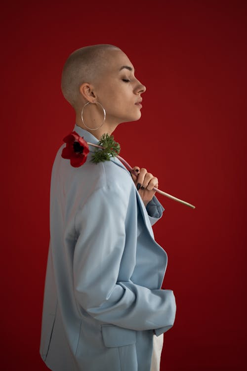 Side view of confident female with short blond hair in stylish clothes standing with red poppy on shoulder in studio against red background