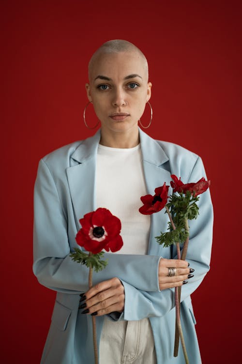 Confident female with short blond hair in stylish clothes standing with crossed arms and holding red poppies in studio against red background