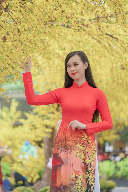 Selective Focus Photo of a Woman in a Red Dress Touching the Yellow Flowers of a Tree
