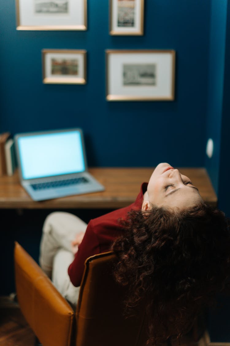 Woman Sitting In An Armchair At The Desk In Front Of A Laptop And Throwing Her Head Back 