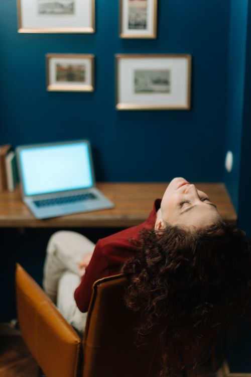Free Woman sitting back in chair at a wooden desk with laptop in a cozy, artistic office space. Stock Photo