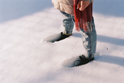 Close-up of a person standing in snow wearing denim and a red scarf, highlighting winter fashion.