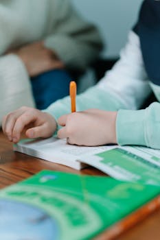 Close-up of a child's hands writing with an orange pen on a book indoors.
