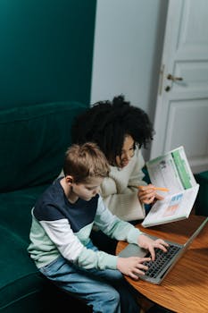 A woman tutors a child with a laptop and book, fostering learning in a cozy setting.