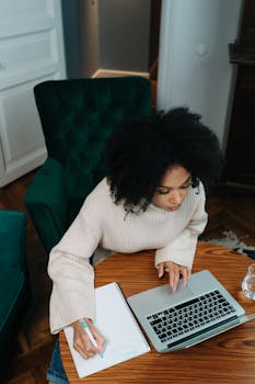 Woman writing notes while using a laptop at a wooden desk, focuses on multitasking and productivity.